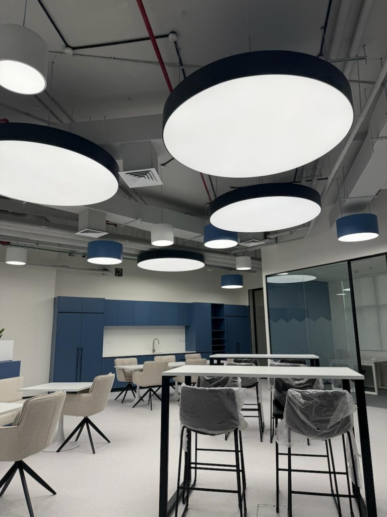 Modern coworking office interior featuring suspended illuminated light boxes with uniform 6500K LED lighting in Dubai Investment Park. The ceiling design creates a professional and productive workspace atmosphere. Modern coworking office interior featuring suspended illuminated light boxes with uniform 6500K LED lighting in Dubai Investment Park. The ceiling design creates a professional and productive workspace atmosphere.