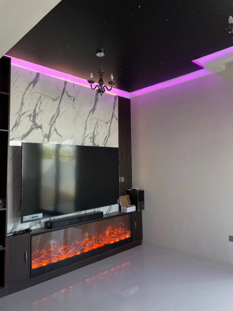 Modern black stretch ceiling with blue RGBW perimeter lighting and integrated micro star effect above a contemporary entertainment wall and fireplace installation in a Dubai villa. Modern black stretch ceiling with blue RGBW perimeter lighting and integrated micro star effect above a contemporary entertainment wall and fireplace installation in a Dubai villa.